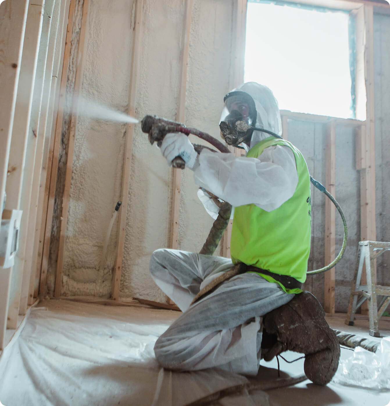 Professional installer in full protective gear applying spray foam insulation to residential wall studs during a home renovation.